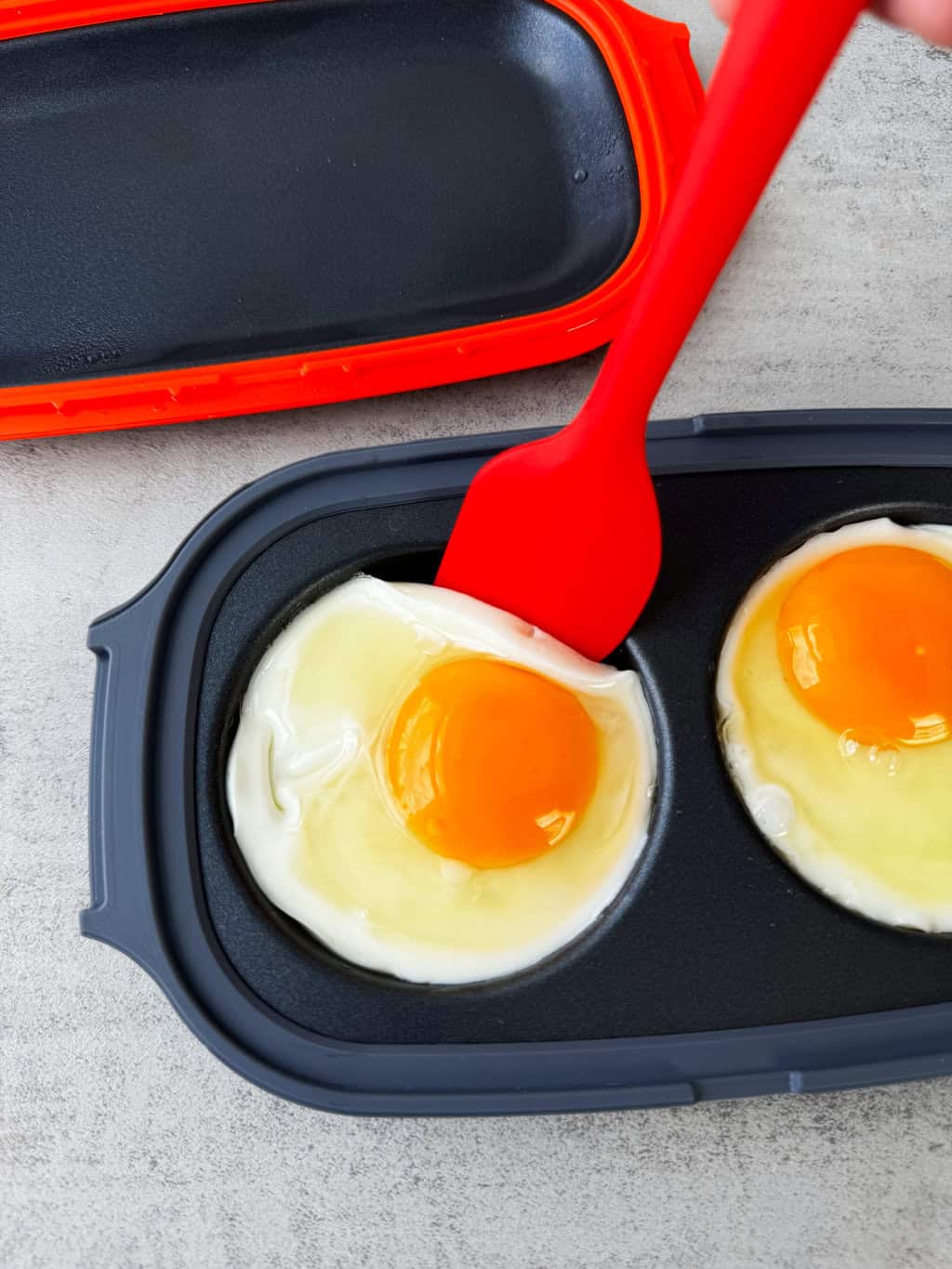 semi cooked eggs in egg maker with silicone spatula lifting an egg to show that it isnt fully cooked yet